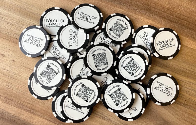 Op de afbeelding: Een verzameling zwart-witte pokerchips met de tekst "Touch of Grace" en "Verwijs een vriend voor $25 korting". De chips hebben een cirkelvormig ontwerp met een zwart-wit gestreepte rand.