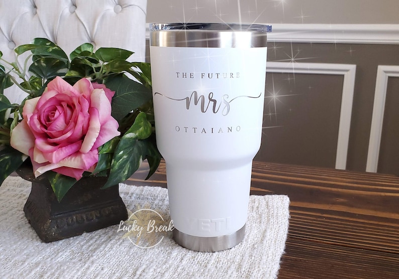 May include: White insulated tumbler with a silver rim and lid. The text "THE FUTURE mrs OTTAIANO" is printed on the front. A pink rose and green foliage are in the background. The tumbler has a silver base.