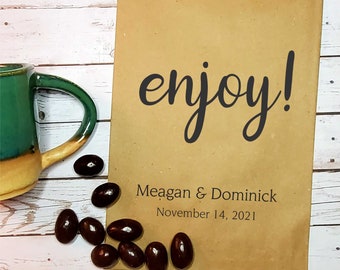 May include: A brown paper bag with the text "enjoy!" in black cursive font. Below the text is the text "Meagan & Dominick" and "November 14, 2021". There are chocolate candies scattered on a wooden surface in front of the bag.