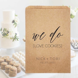 May include: A brown paper bag with the text "we do (love cookies)", "Nick + Tori", and the date "03.27.2021" printed in black. A white tiered wedding cake with floral decorations is in the background.