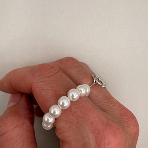 May include: A ring featuring a row of white, round pearls. The pearls are strung together and attached to a silver-colored band. The ring is worn on a finger.