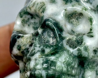 May include: A green and white agate skull sculpture. The skull is carved in detail, with visible eye sockets, a nose, and teeth. The agate is a natural stone with a swirling pattern of green and white.