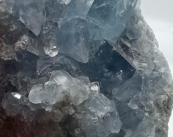 May include: A large, blue celestite crystal cluster with a rough, natural surface. The crystal is composed of many smaller, faceted crystals that are clustered together. The crystal has a light blue color with a slight translucence.