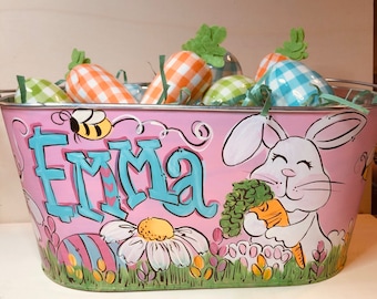 May include: Pink metal Easter basket with the name "EMMA" in blue letters. The basket features a painted bunny, bee, and flowers. It is filled with carrot and egg-shaped decorations.