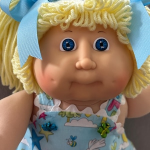May include: A vintage doll with blonde yarn hair styled in pigtails, each tied with a light blue ribbon. The doll has blue eyes and rosy cheeks. It wears a light blue dress patterned with rainbows, bees, and stars.