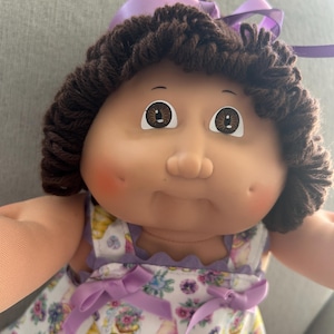 May include: A vintage doll with brown yarn hair and a purple ribbon. The doll has brown eyes and rosy cheeks. It is wearing a white dress with a floral and gnome print, and purple ribbon accents.