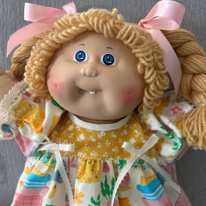 May include: A vintage doll with blonde yarn hair styled in pigtails, each tied with a pink ribbon. The doll has blue eyes, rosy cheeks, and a small gap-toothed smile. It wears a dress with a yellow bodice and a white skirt patterned with colorful spring-themed graphics.