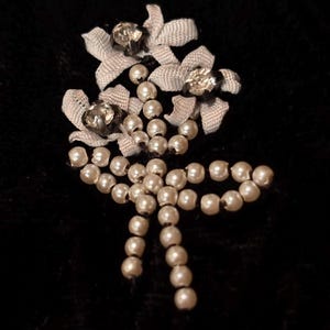 May include: A vintage brooch with a floral design, featuring faux pearls and ribbon details. The brooch has three ribbon flowers, each with a sparkling rhinestone. The design includes a cascade of pearls and a bow.