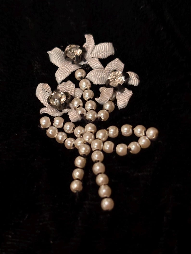 May include: A vintage brooch with a floral design, featuring faux pearls and ribbon details. The brooch has three ribbon flowers, each with a sparkling rhinestone. The design includes a cascade of pearls and a bow.