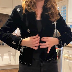 May include: A black velvet jacket with white floral and beaded detailing. The jacket features a scalloped hem and cuffs. The sleeves are long and the jacket is open at the front. It appears to be a formal or evening wear garment.