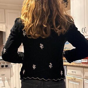 May include: Black velvet jacket with long sleeves and a scalloped hem. The jacket is decorated with embroidered floral designs in white thread. The garment is paired with dark wash denim jeans.