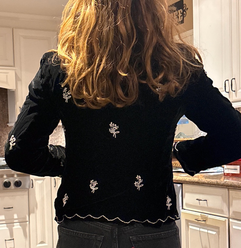 May include: Black velvet jacket with long sleeves and a scalloped hem. The jacket is decorated with embroidered floral designs in white thread. The garment is paired with dark wash denim jeans.