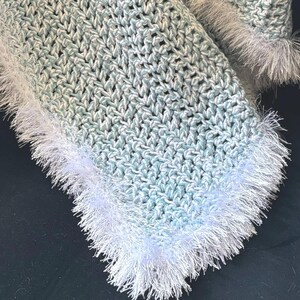 May include: Crocheted blanket in shades of light blue and white, with a textured stitch pattern. The edges are trimmed with a fluffy, white fringe. The blanket is handmade and appears soft and cozy.