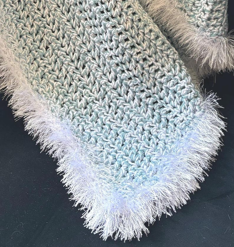 May include: Crocheted blanket in shades of light blue and white, with a textured stitch pattern. The edges are trimmed with a fluffy, white fringe. The blanket is handmade and appears soft and cozy.