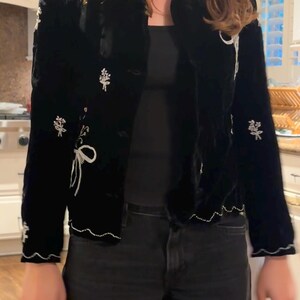 May include: Black velvet jacket with white floral embroidery and a scalloped hem. The jacket has a button-up front and a classic collar. The floral embroidery is detailed with white thread, adding a delicate touch to the garment.