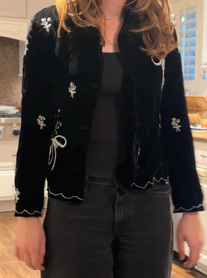 May include: Black velvet jacket with white floral embroidery and a scalloped hem. The jacket has a button-up front and a classic collar. The floral embroidery is detailed with white thread, adding a delicate touch to the garment.