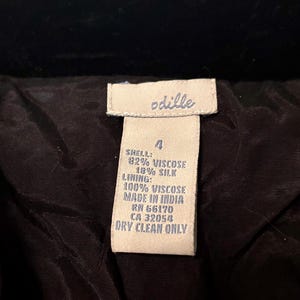 May include: A close-up of a clothing label with the brand name "odille" in white text. The label details the shell composition as 82% viscose and 18% silk, with a 100% viscose lining. It states the item was made in India and requires dry cleaning.