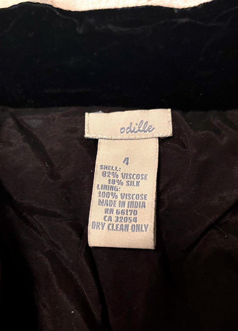 May include: A close-up of a clothing label with the brand name "odille" in white text. The label details the shell composition as 82% viscose and 18% silk, with a 100% viscose lining. It states the item was made in India and requires dry cleaning.