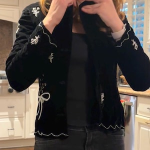 May include: Black velvet jacket with white embroidered floral and bow details. The jacket features a classic design with a notched collar and button closure. The cuffs and hem are trimmed with a delicate white beaded edge.