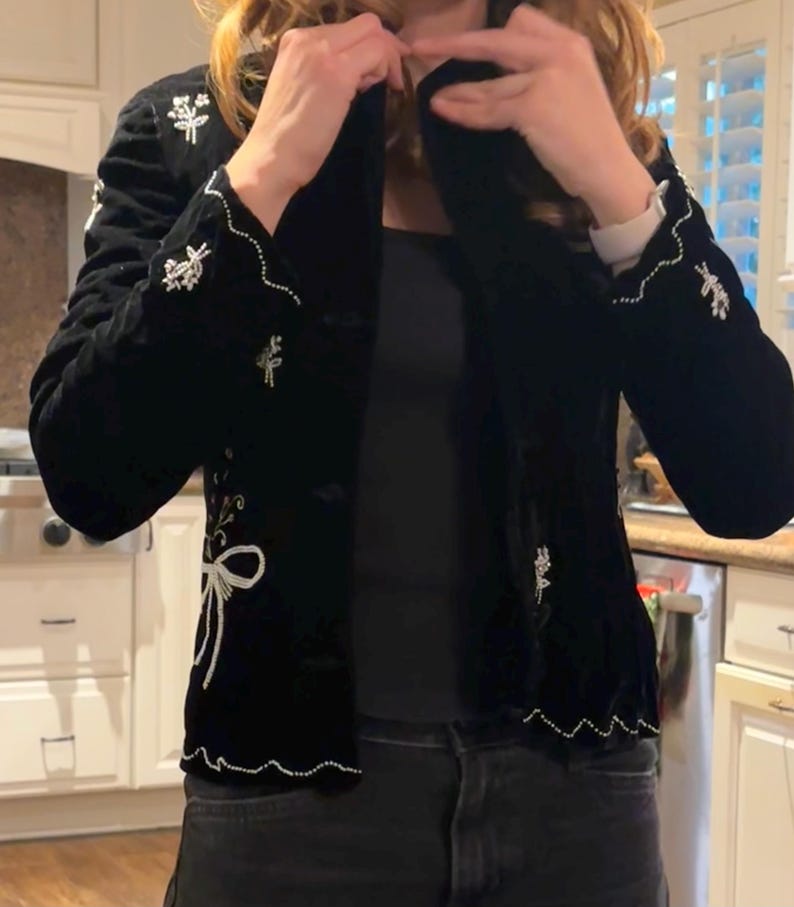 May include: Black velvet jacket with white embroidered floral and bow details. The jacket features a classic design with a notched collar and button closure. The cuffs and hem are trimmed with a delicate white beaded edge.