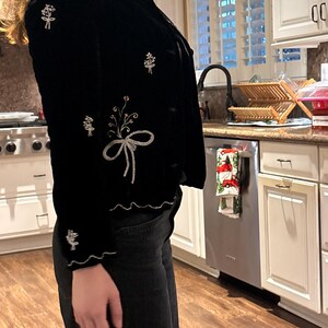 May include: Black velvet jacket with floral embroidery in white, pink, and silver thread. The jacket features a scalloped hem and cuffs, and a bow design on the front. The jacket has a button closure and a ruffled collar.
