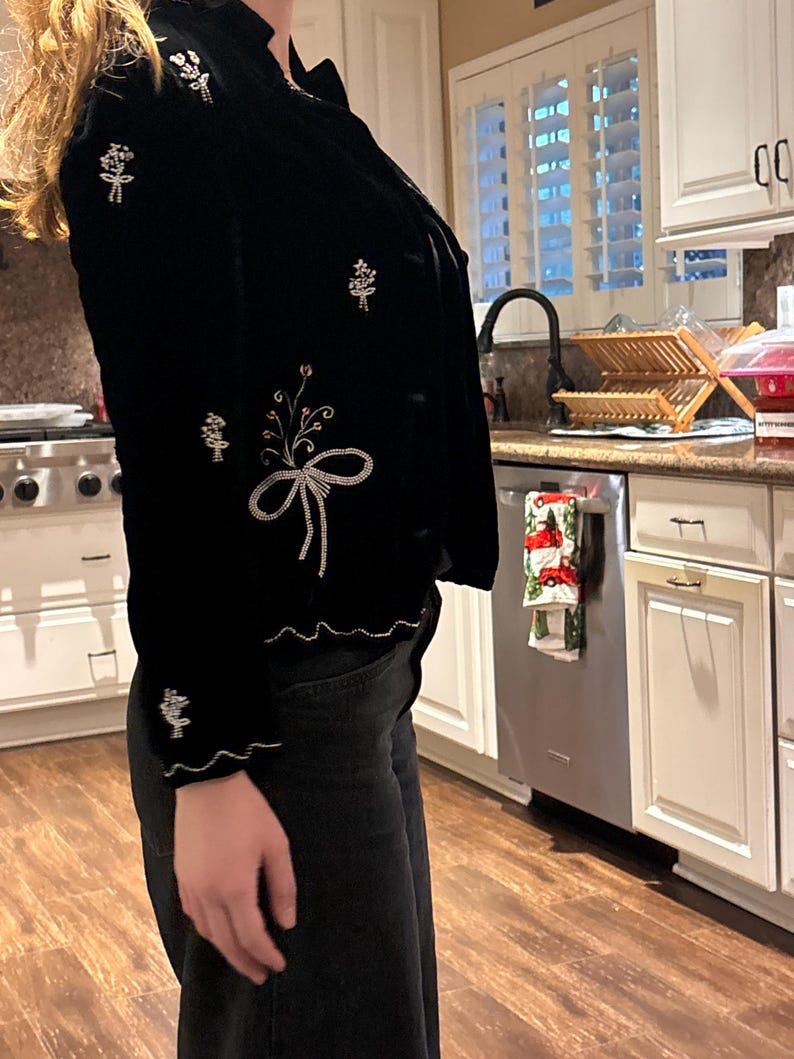 May include: Black velvet jacket with floral embroidery in white, pink, and silver thread. The jacket features a scalloped hem and cuffs, and a bow design on the front. The jacket has a button closure and a ruffled collar.