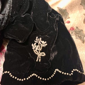 May include: A close-up of a black velvet garment with a decorative white floral brooch. The edge is trimmed with small, white, pearl-like beads. The velvet has a soft, textured appearance.