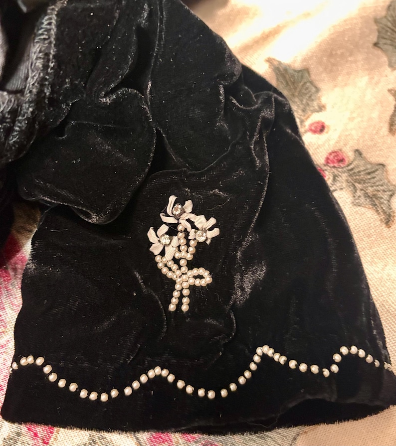 May include: A close-up of a black velvet garment with a decorative white floral brooch. The edge is trimmed with small, white, pearl-like beads. The velvet has a soft, textured appearance.