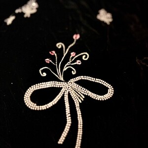 May include: A detailed shot of black fabric with an embroidered design. The design includes a bow with trailing ribbons and a floral element with pink accents, all in a light, shimmering colour. The fabric has a velvety texture.