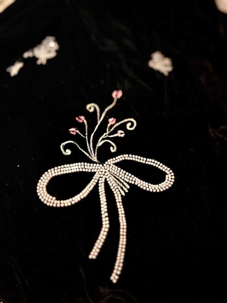 May include: A detailed shot of black fabric with an embroidered design. The design includes a bow with trailing ribbons and a floral element with pink accents, all in a light, shimmering colour. The fabric has a velvety texture.