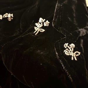 May include: A close-up of a black velvet jacket adorned with floral brooches. The jacket is embellished with several clusters of small, white, and silver-toned floral brooches. The fabric has a soft, textured appearance.