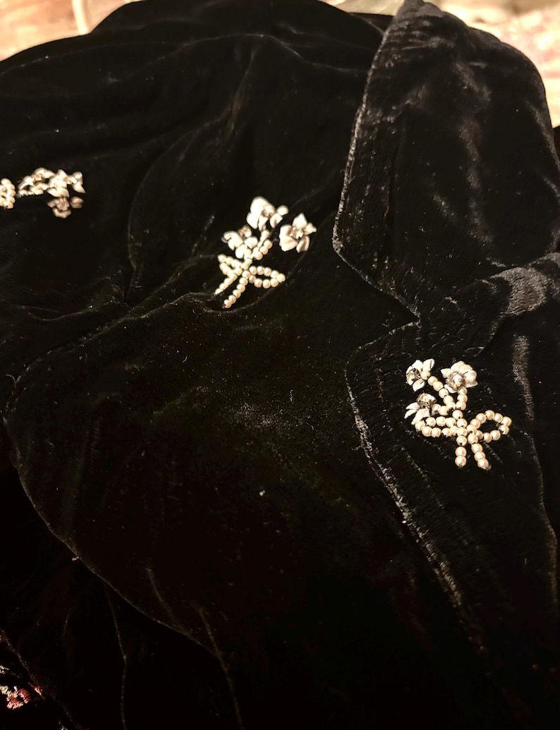 May include: A close-up of a black velvet jacket adorned with floral brooches. The jacket is embellished with several clusters of small, white, and silver-toned floral brooches. The fabric has a soft, textured appearance.