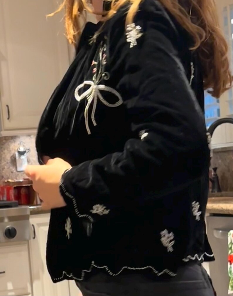 May include: Black velvet jacket with white floral and bow embroidery. The jacket features a scalloped hem and detailed embellishments. A stylish outerwear piece.