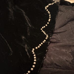 May include: Close-up of black velvet fabric with a beaded trim. The trim is a string of small, round, white beads that follows the edge of the velvet. The fabric has a soft, textured appearance.