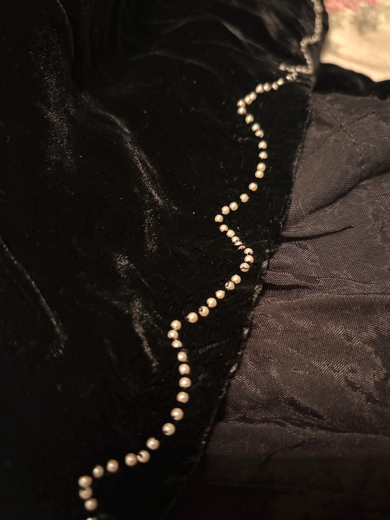 May include: Close-up of black velvet fabric with a beaded trim. The trim is a string of small, round, white beads that follows the edge of the velvet. The fabric has a soft, textured appearance.