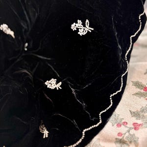 May include: A black velvet shawl with floral rhinestone embellishments. The shawl has a scalloped edge trimmed with small white beads. The floral designs are clustered in various areas of the shawl.