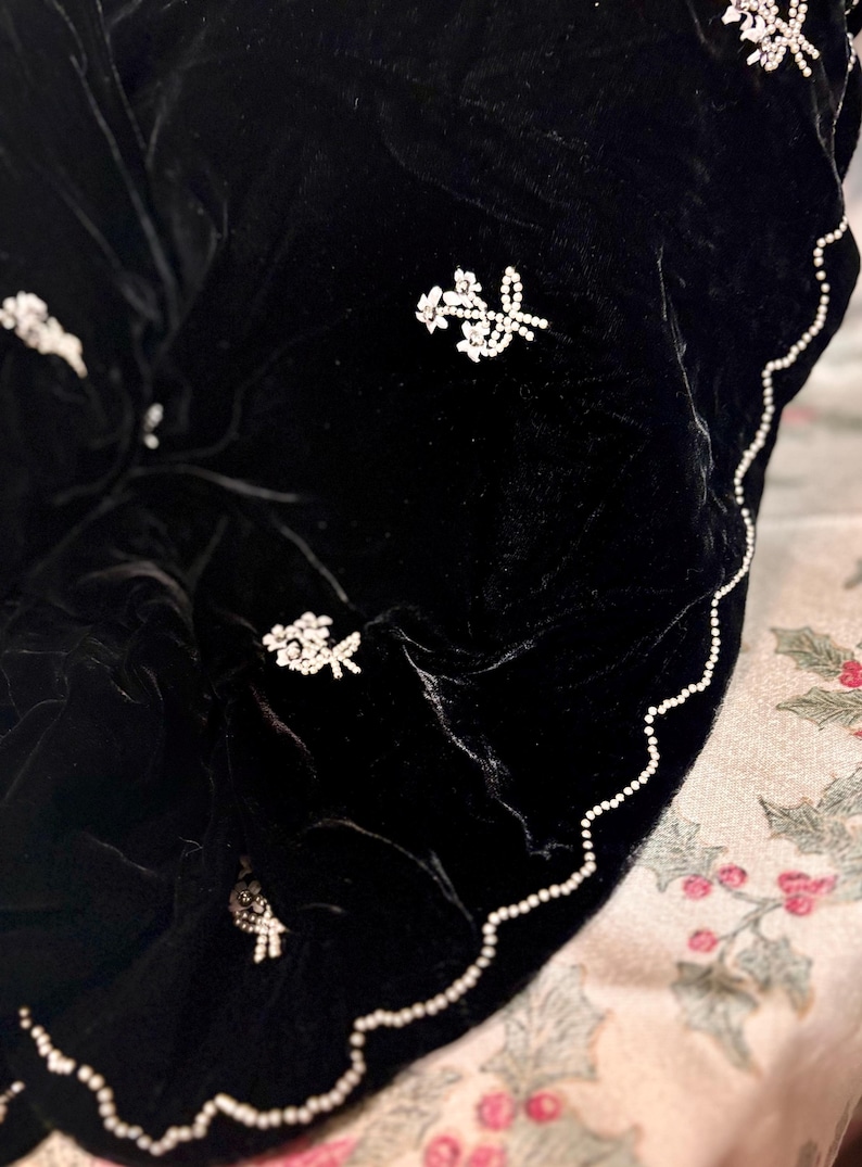 May include: A black velvet shawl with floral rhinestone embellishments. The shawl has a scalloped edge trimmed with small white beads. The floral designs are clustered in various areas of the shawl.