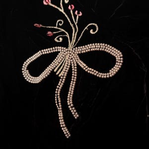 May include: Close-up of black velvet fabric with an embroidered design. The design features a bow made of small, pearl-like beads, with a floral accent of pink buds and delicate stems.