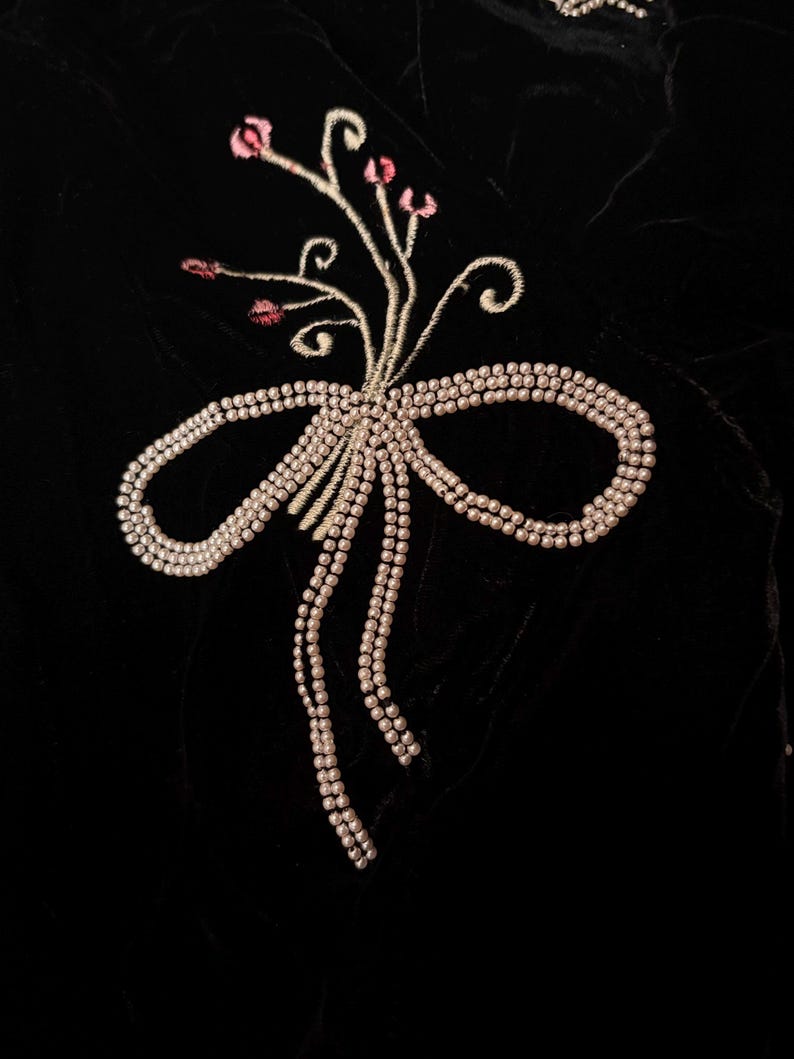 May include: Close-up of black velvet fabric with an embroidered design. The design features a bow made of small, pearl-like beads, with a floral accent of pink buds and delicate stems.