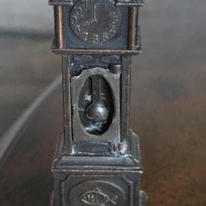 May include: A miniature antique-style clock in a dark metallic finish. The clock features a detailed face with Roman numerals and decorative elements. The base has an oval emblem with a fish design.