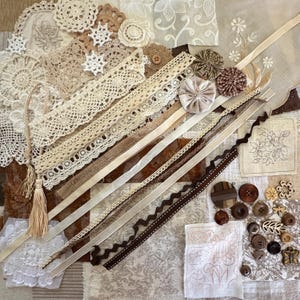 May include: A selection of vintage craft materials in cream, beige, and brown tones. The image shows lace trim, ribbons, buttons, doilies, and fabric patches, suitable for scrapbooking and other craft projects.