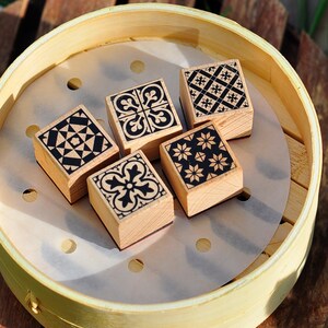 May include: Four wooden stamps with different black and white geometric patterns. The stamps are arranged in a circle on a white paper liner inside a round bamboo steamer.