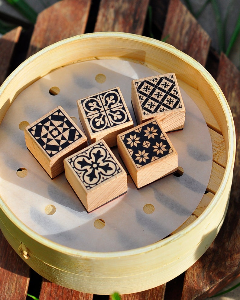 May include: Four wooden stamps with different black and white geometric patterns. The stamps are arranged in a circle on a white paper liner inside a round bamboo steamer.