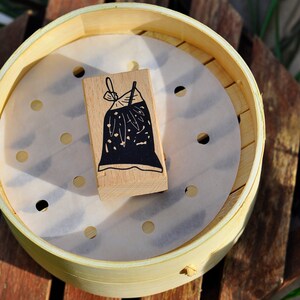 May include: A wooden block with a black and white illustration of a bag with a bow on top. The block is sitting on a white paper liner inside a round, light brown bamboo steamer.