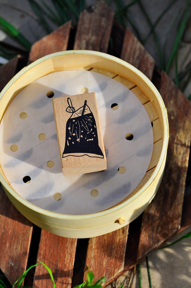 May include: A wooden block with a black and white illustration of a bag with a bow on top. The block is sitting on a white paper liner inside a round, light brown bamboo steamer.