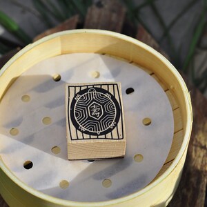May include: A wooden stamp with a black and white design of a turtle with the Chinese character for "happiness" inside. The stamp is sitting inside a bamboo steamer.