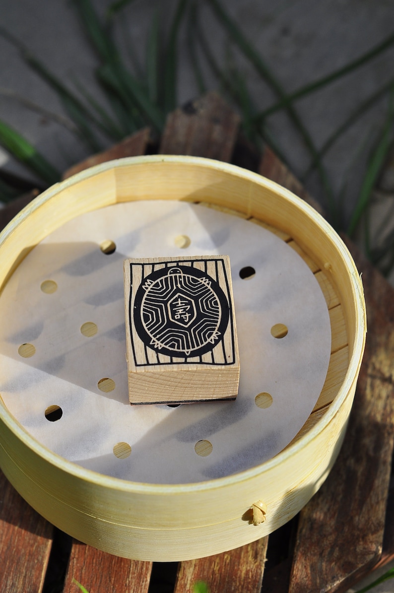 May include: A wooden stamp with a black and white design of a turtle with the Chinese character for "happiness" inside. The stamp is sitting inside a bamboo steamer.