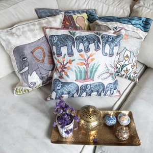 May include: A set of four decorative throw pillows with embroidered elephant designs. The pillows are in shades of white, gray, blue, and green. The pillows are arranged on a white couch.