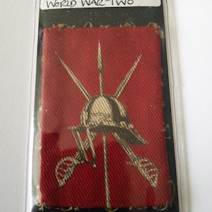 May include: A red fabric patch with a gold embroidered design of a helmet with crossed swords. The patch has the text "Central Military District East Central District (Eastern Command) World War Two" written on a white label.