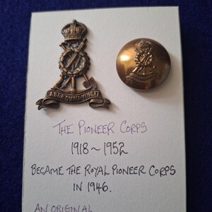 May include: A display of two vintage Pioneer Corps insignia. One is a detailed emblem with a crown and crossed tools, the other a button. The card states "The Pioneer Corps 1918-1952" and "Became the Royal Pioneer Corps in 1946."